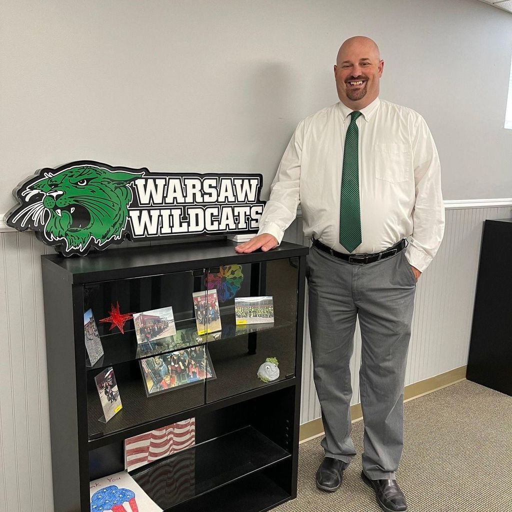 Man in a green tie standing next to a book shelf with photos on it